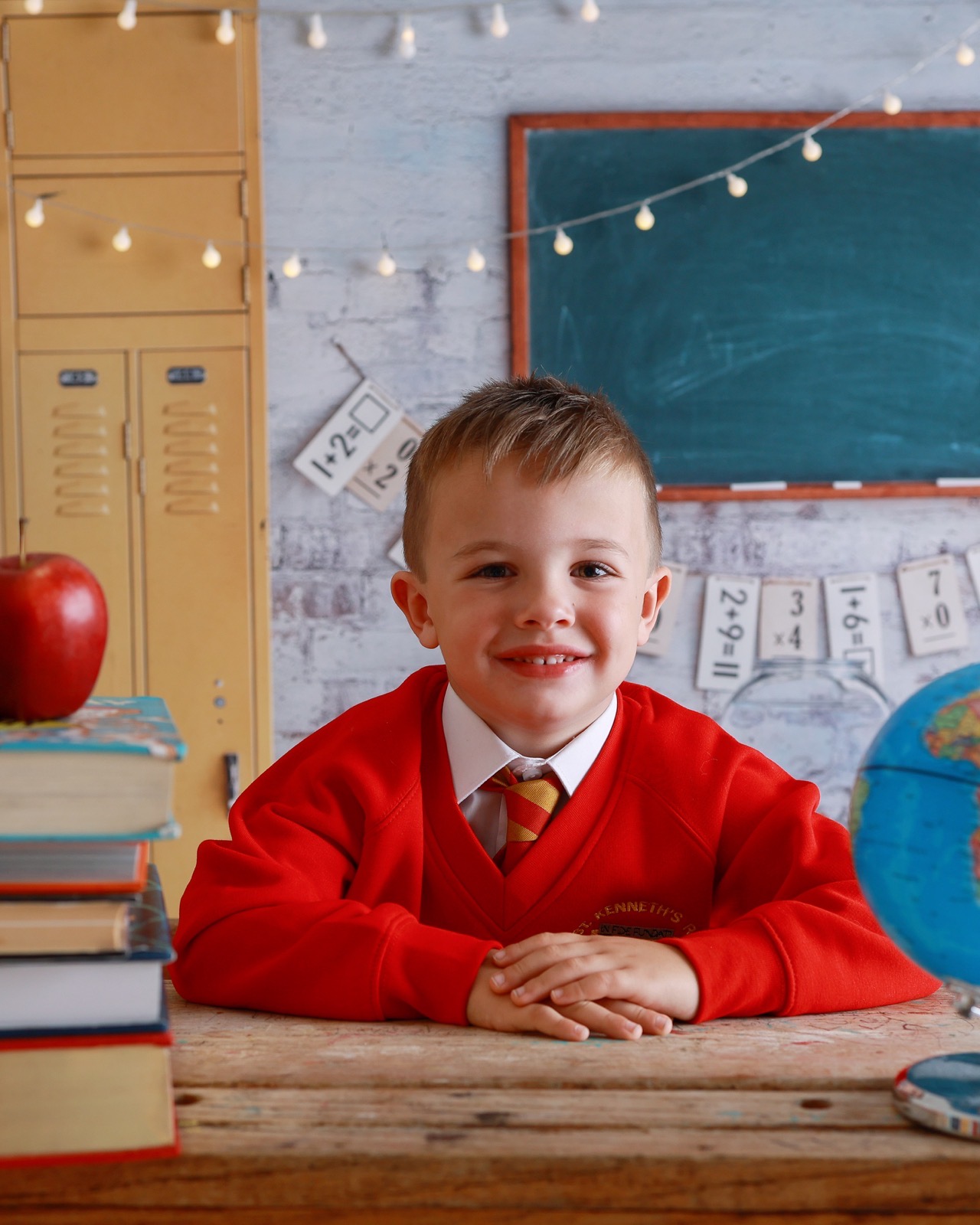 Close-up portrait of a smiling boy in a red jumper and striped tie, sitting at a wooden desk
