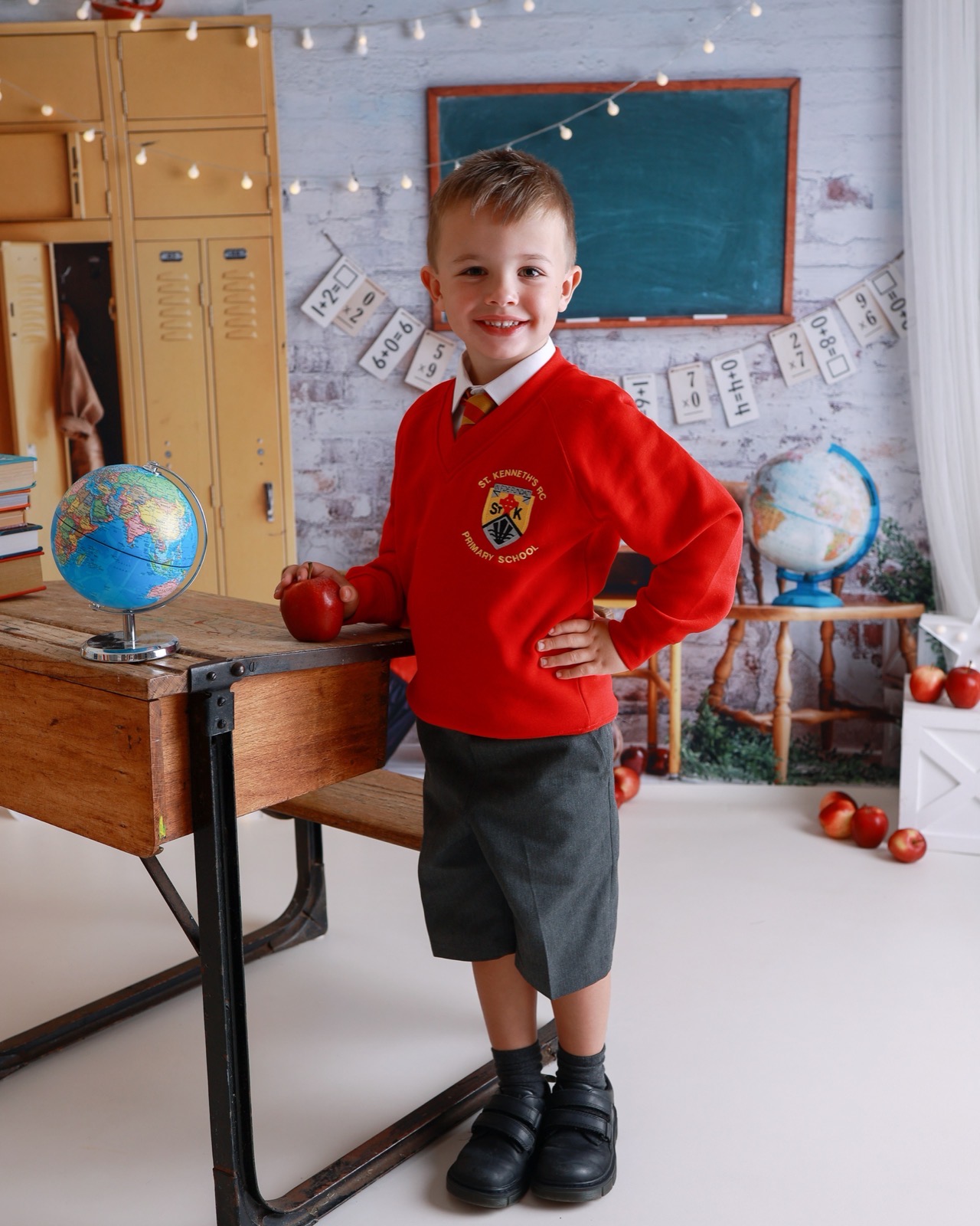 Boy standing by a desk holding a red apple, red school jumper and grey shorts, globe and books nearby
