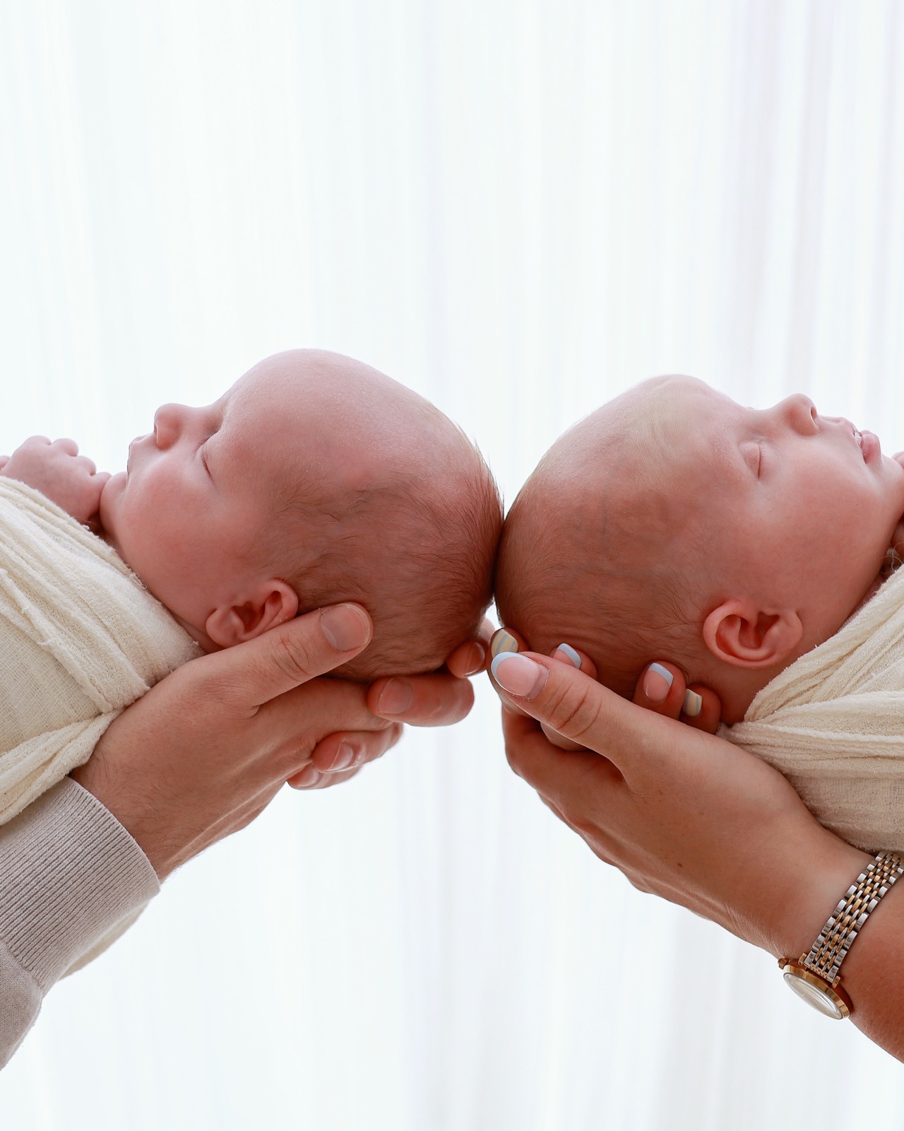 Newborn twins lying head to head, eyes closed, held gently by their parents' hands on either side
