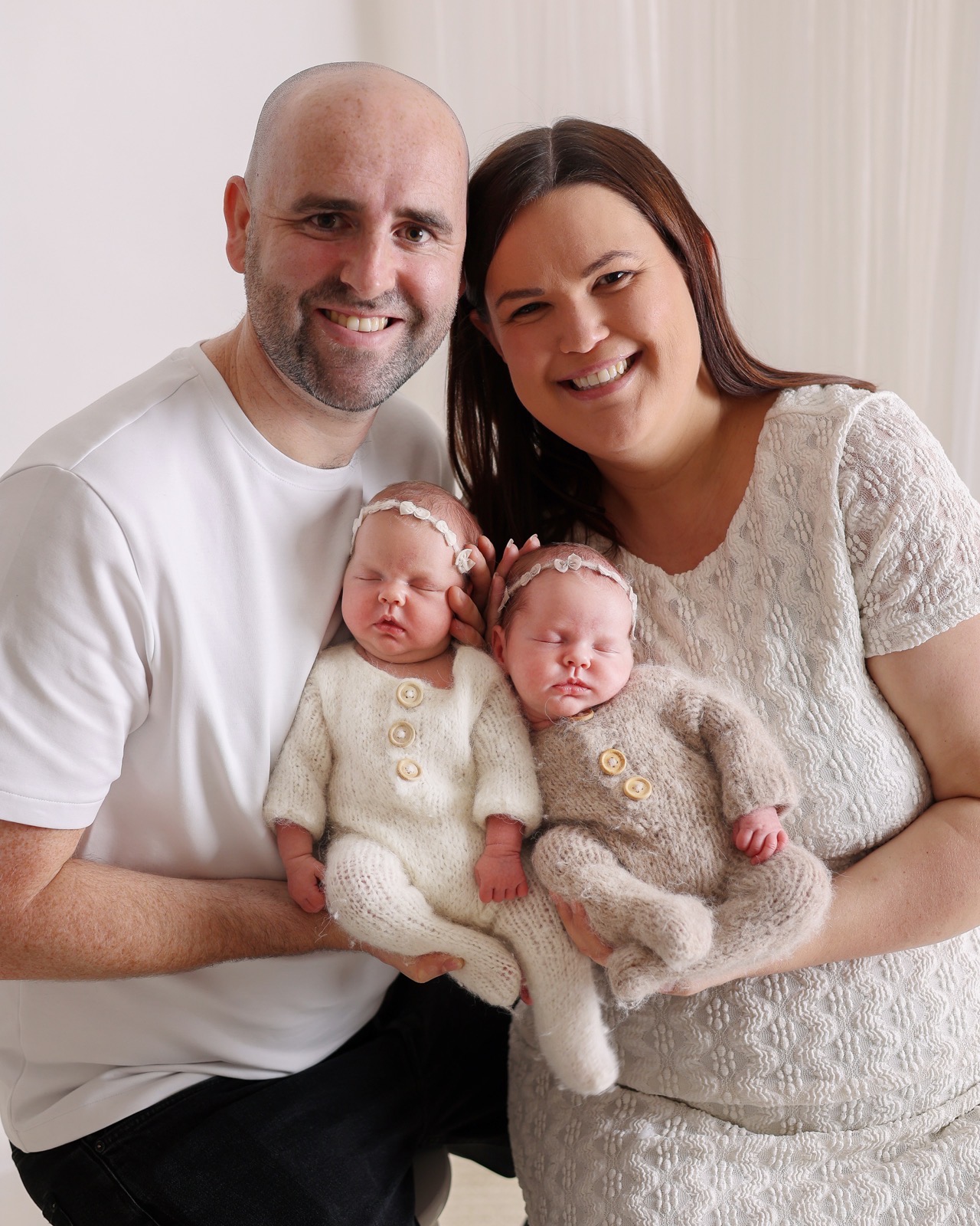 Mum and dad smiling, each cradling one of their newborn twins in matching cream knitted rompers