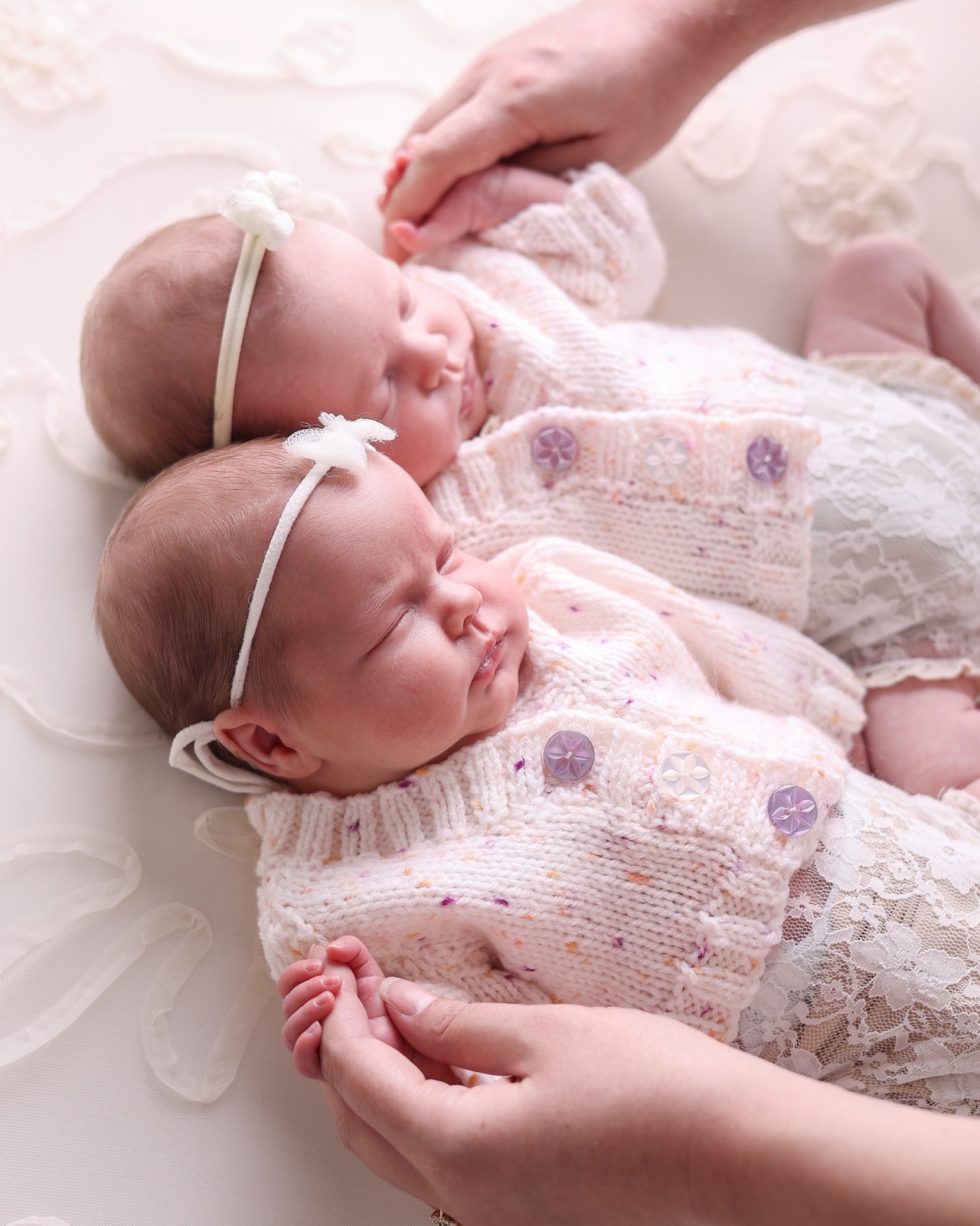 Newborn twin girls lying side by side in matching lace outfits and bow headbands, snuggled together