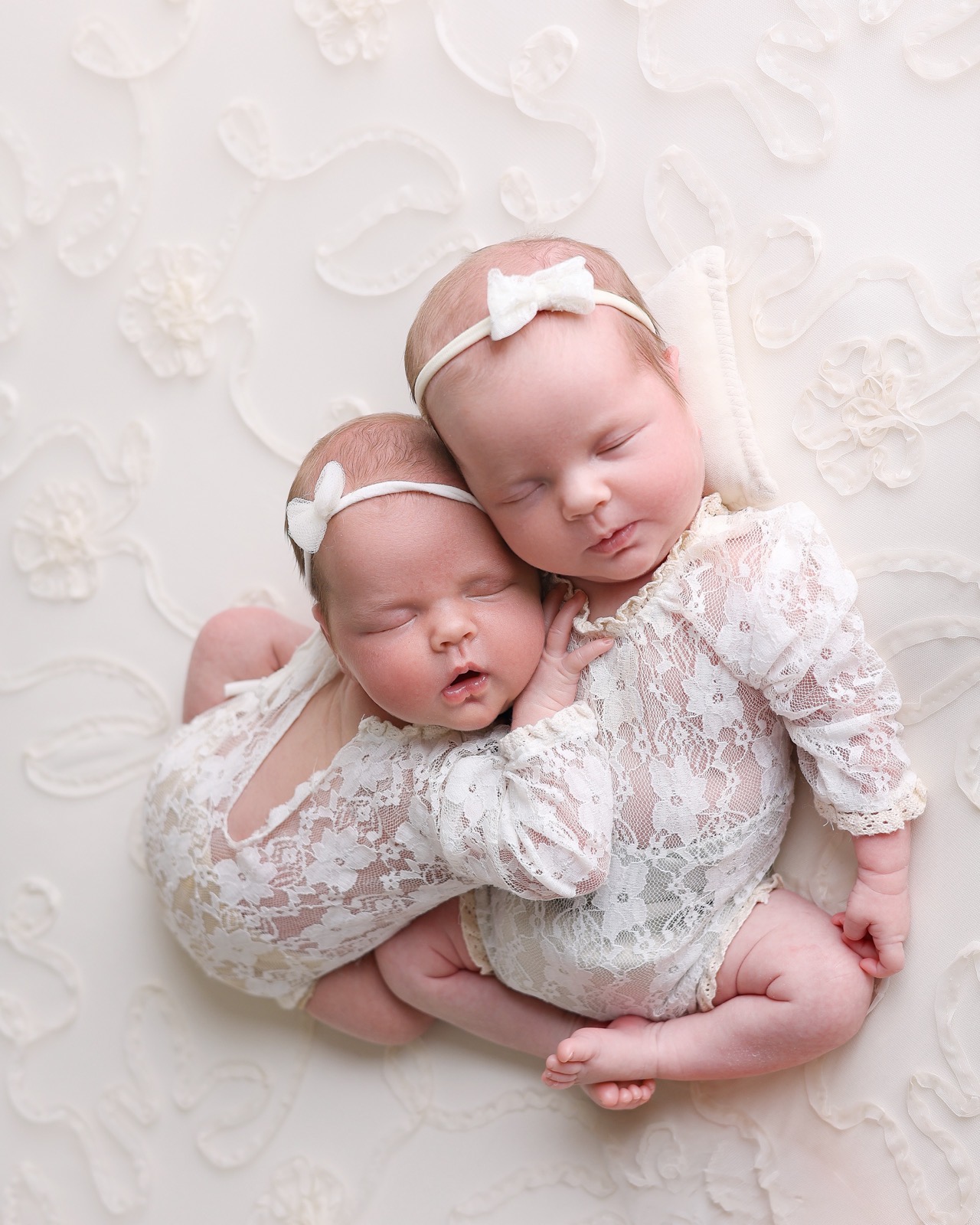 Newborn twin girls cuddled together in cream lace rompers, one resting her head on her sister's chest