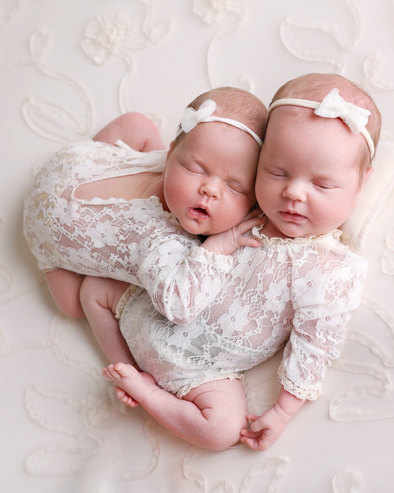 Newborn twin girls lying face to face on a lace blanket, matching bow headbands, peacefully asleep