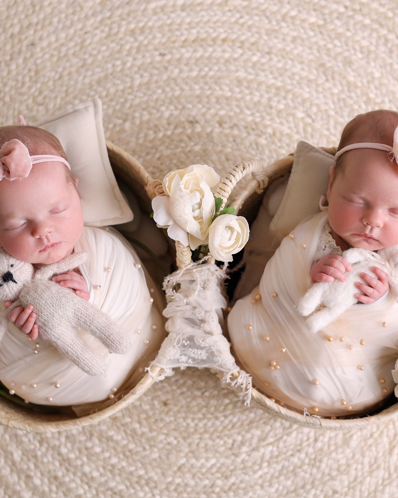 Newborn twin girls asleep side by side in matching woven baskets, each cuddling a knitted bunny toy