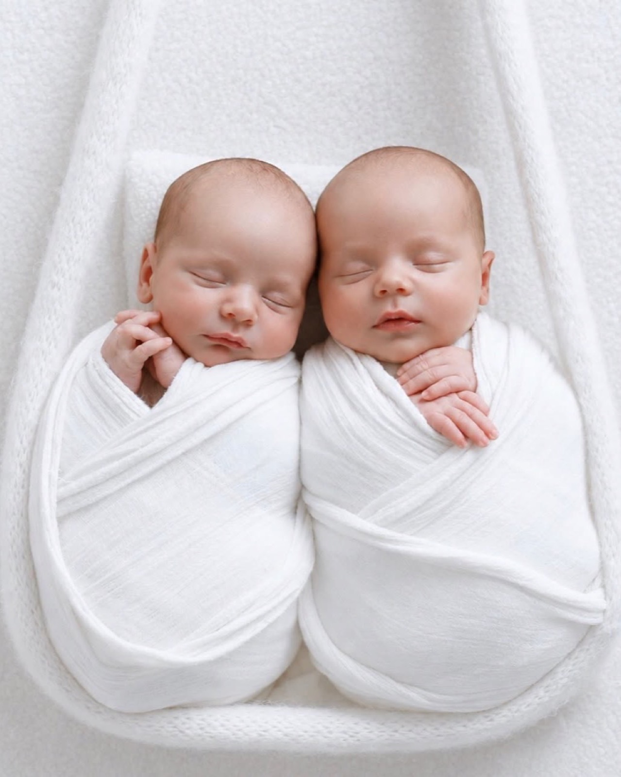 Newborn twin boys swaddled in white wraps, sleeping side by side with heads touching