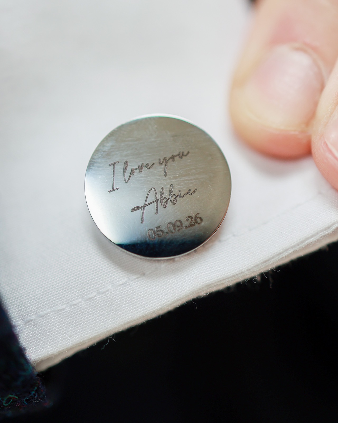 Close-up of an engraved silver cufflink with a personal message and wedding date, held between a groomsman's fingers