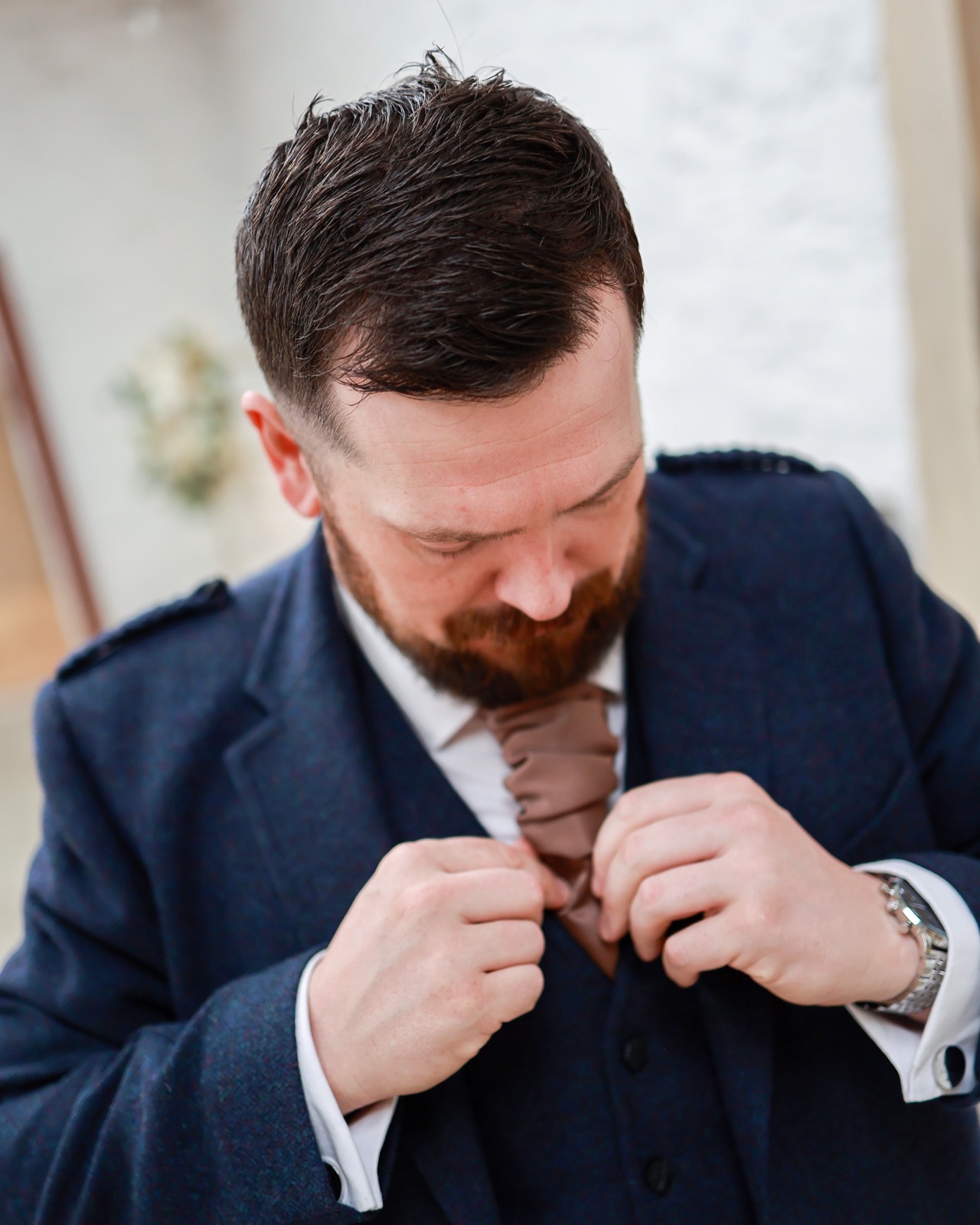 Groom adjusting his brown tie, navy tweed three-piece suit with a tartan-trimmed epaulette