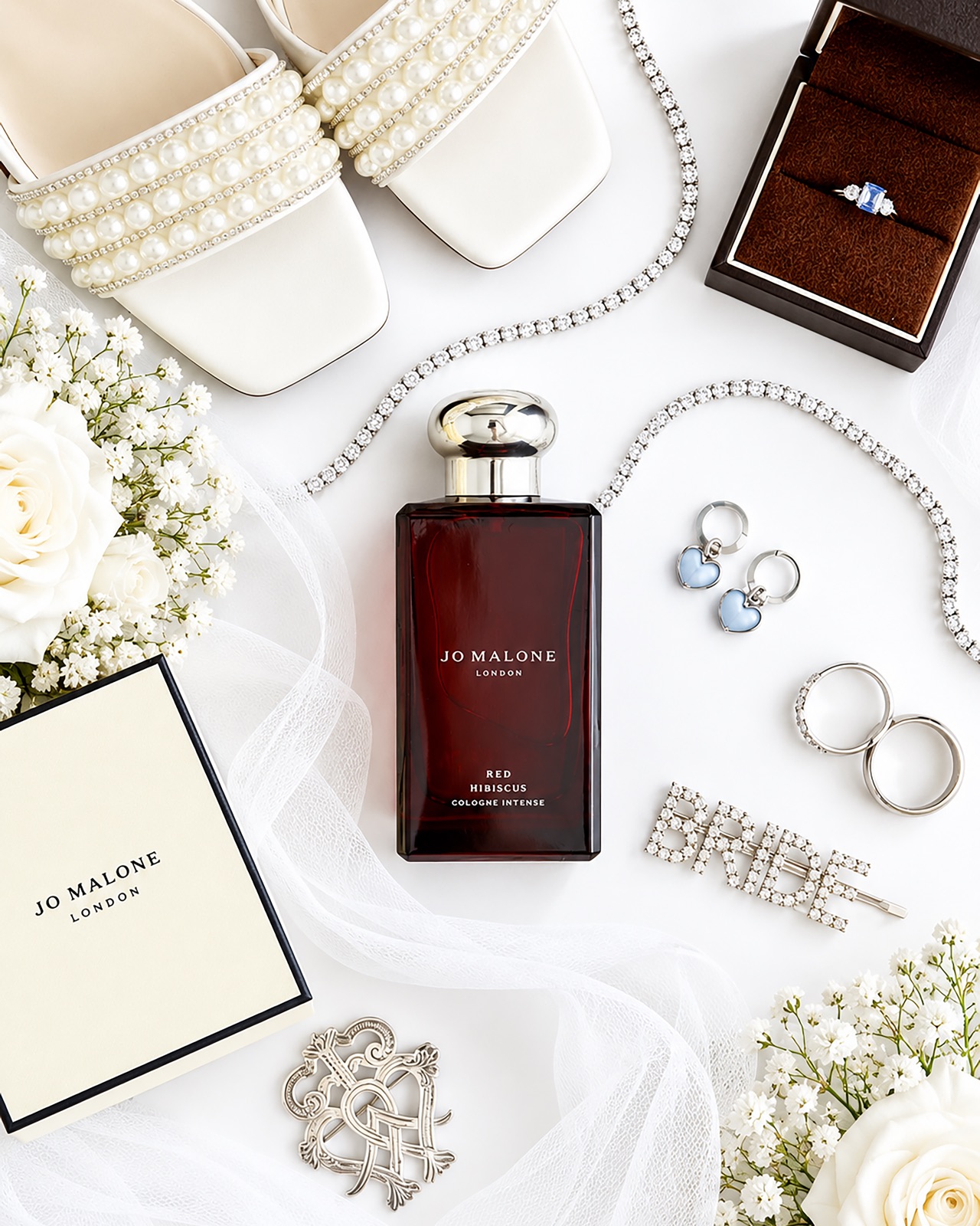 Bridal flat-lay of pearl-embellished shoes, a diamante necklace, engagement ring box, perfume, earrings, wedding bands, a 'BRIDE' hair clip, white roses and a veil