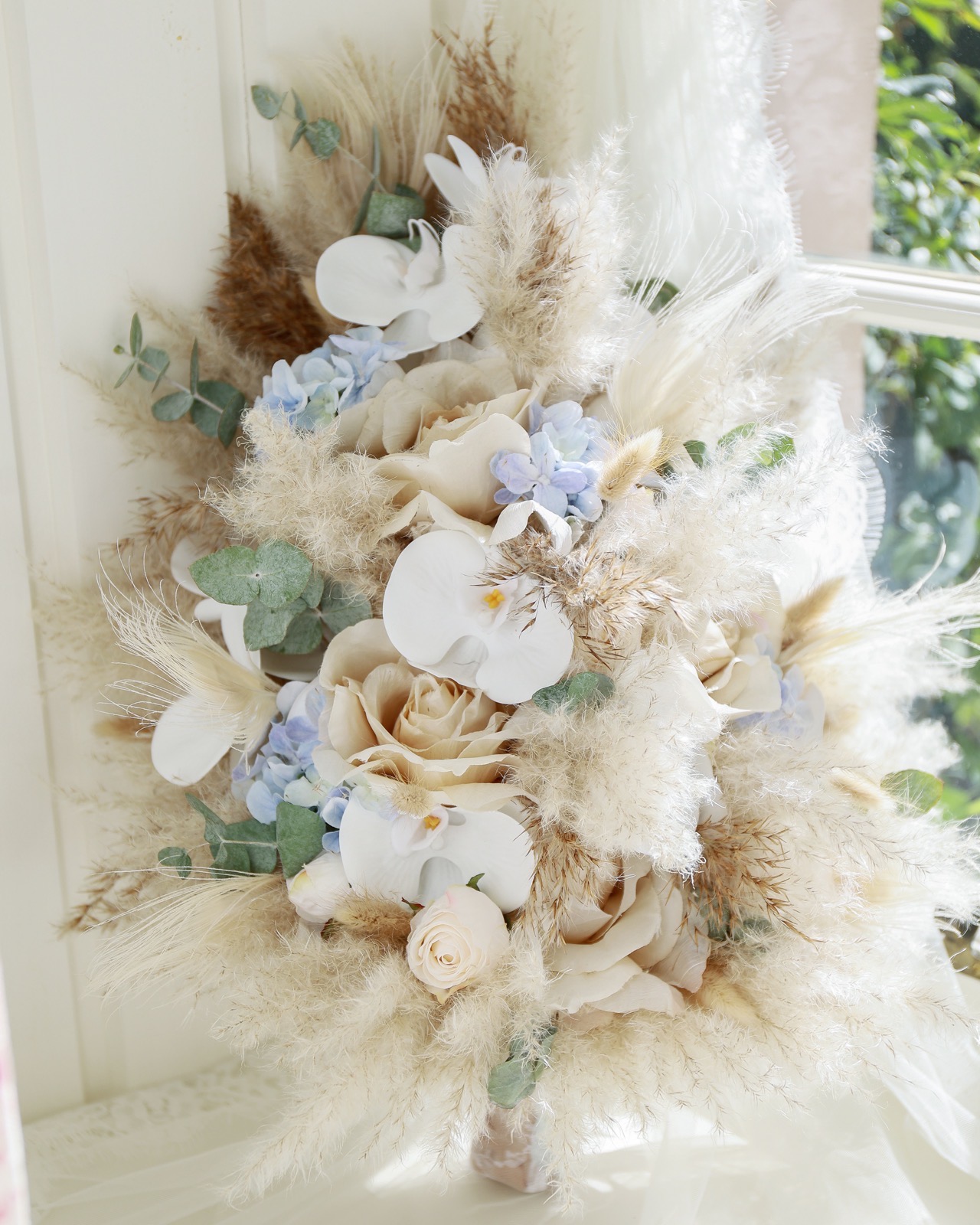 Bridal bouquet of cream roses, white orchids, blue hydrangea, dried pampas grass and eucalyptus by a window
