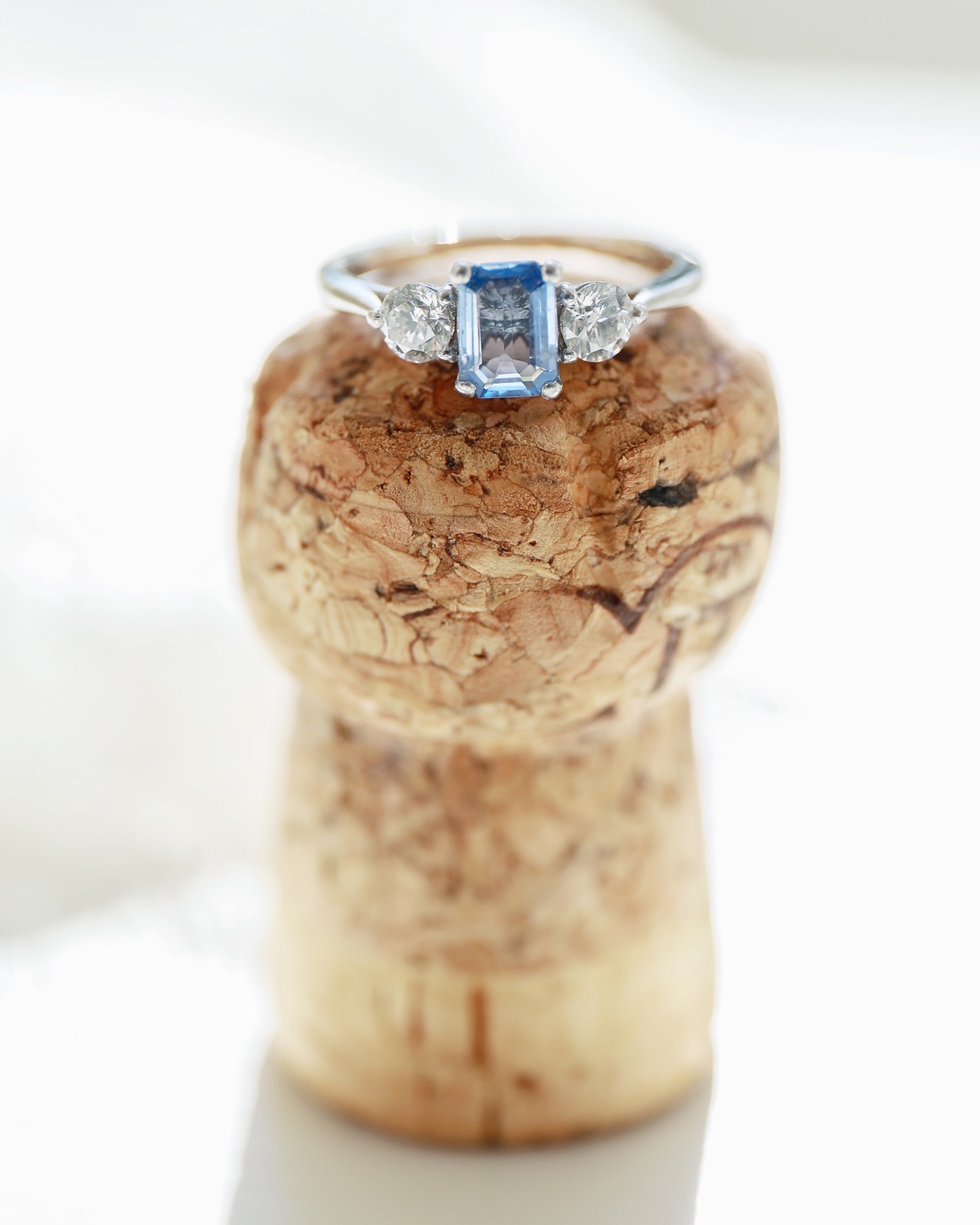 Close-up of a sapphire and diamond three-stone engagement ring balanced on a champagne cork