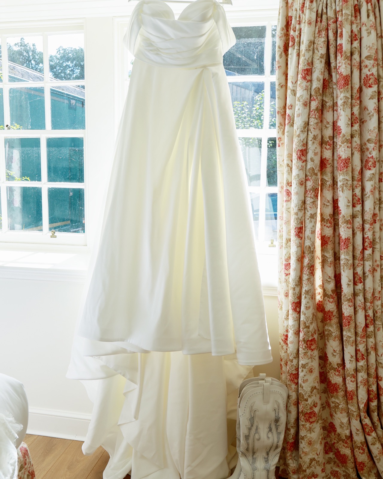 Strapless ivory wedding dress hanging in a window with rhinestone-embroidered boots on the floor beneath