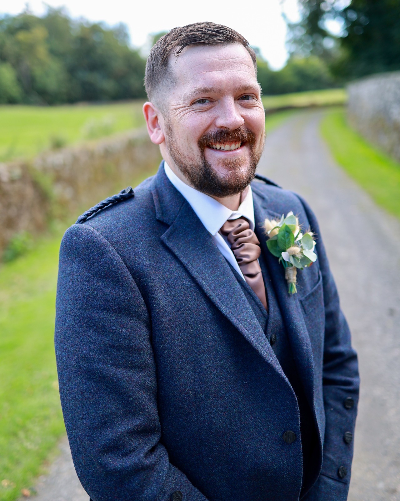 Close-up portrait of a groomsman with a beard, navy tweed jacket and a satin cravat-style tie