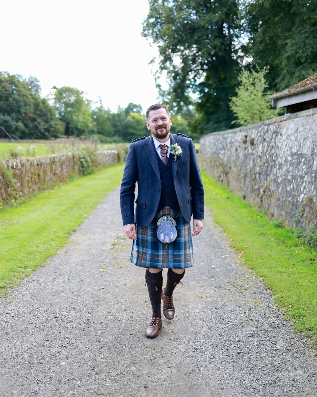 Groomsman walking down a gravel path in a navy tweed jacket and tartan kilt