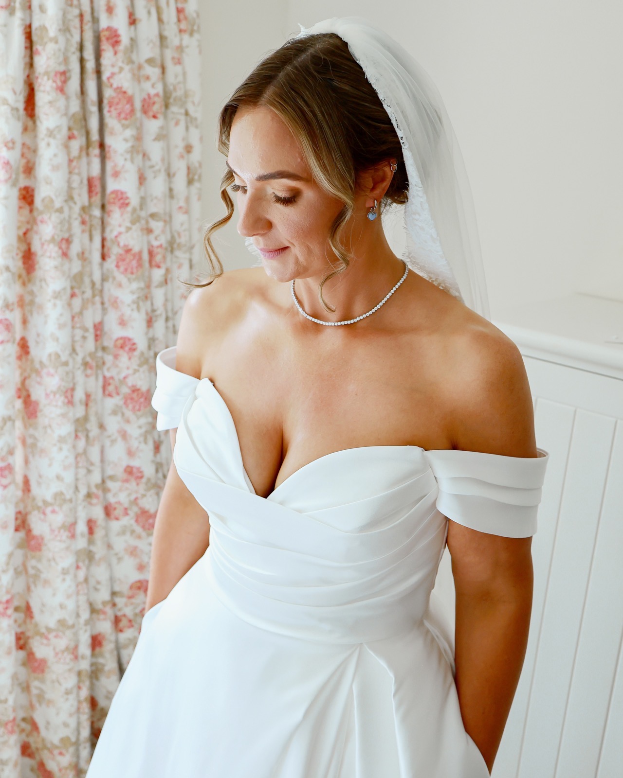 Close-up of a bride in an off-the-shoulder wedding dress and veil, looking down, pearl choker necklace