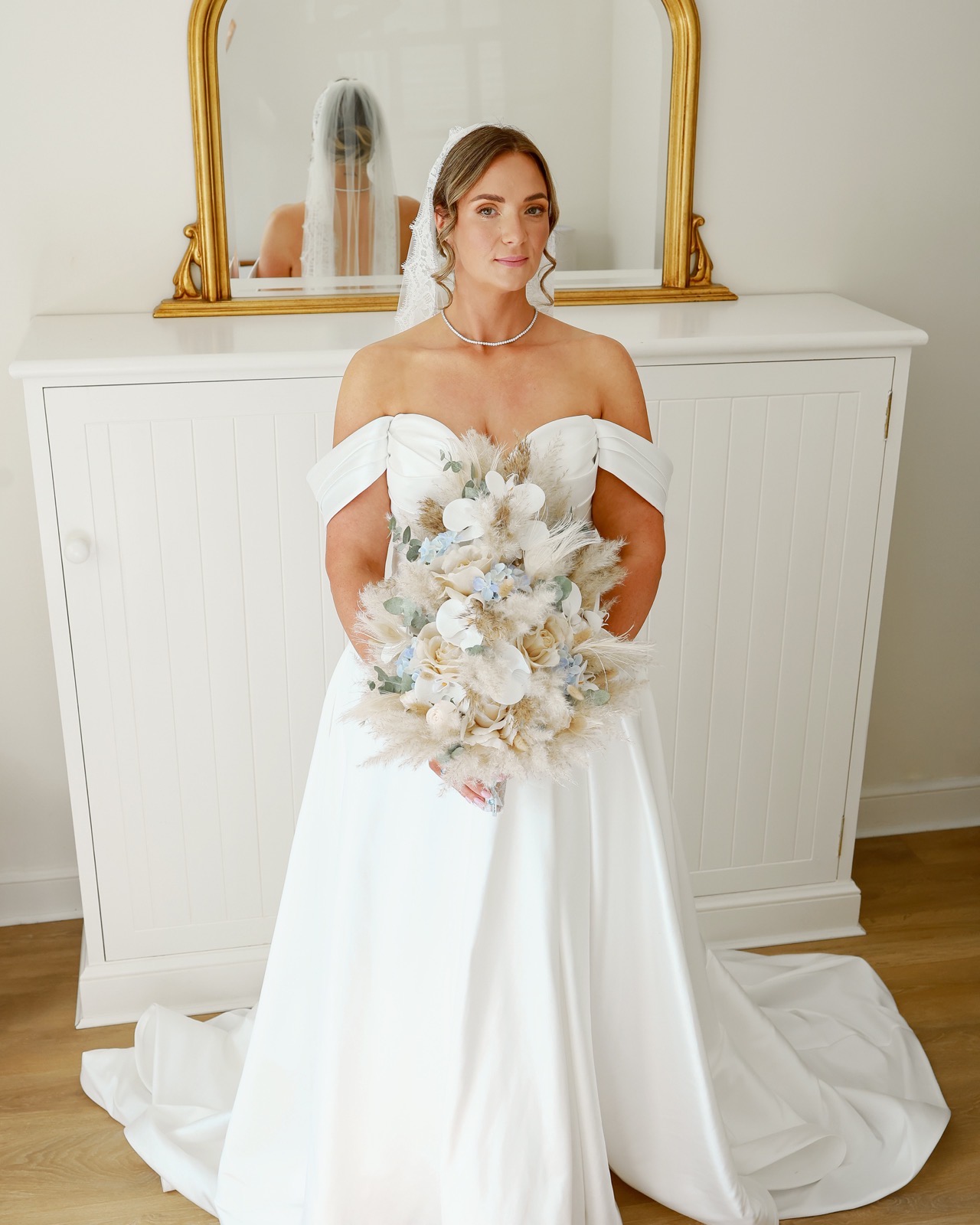 Bride standing in front of a gilt mirror and dresser, holding her bouquet, looking softly at the camera