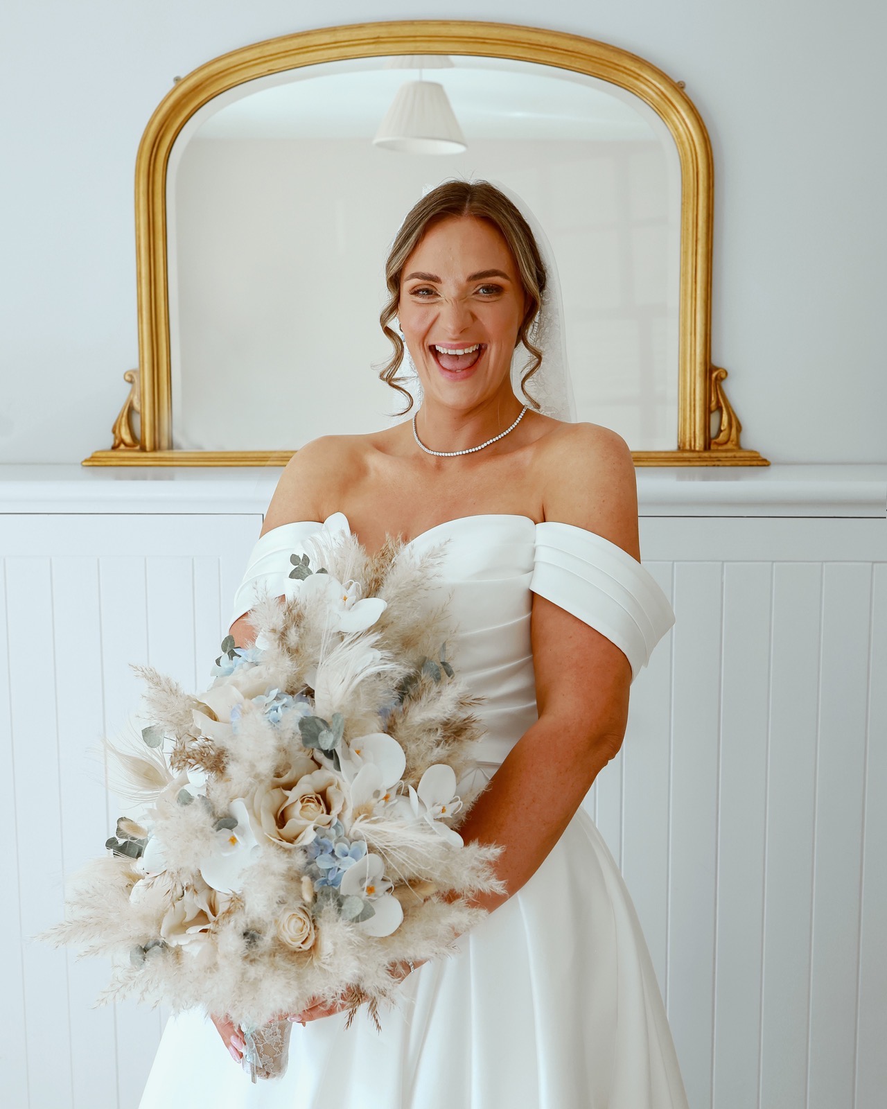 Bride laughing joyfully in front of a gilt mirror, holding her bouquet