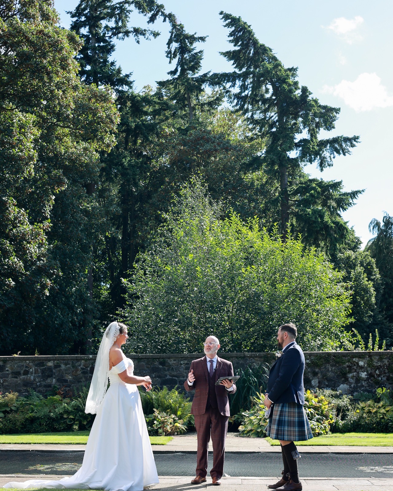 Outdoor wedding ceremony, bride and groom facing the celebrant in a walled garden