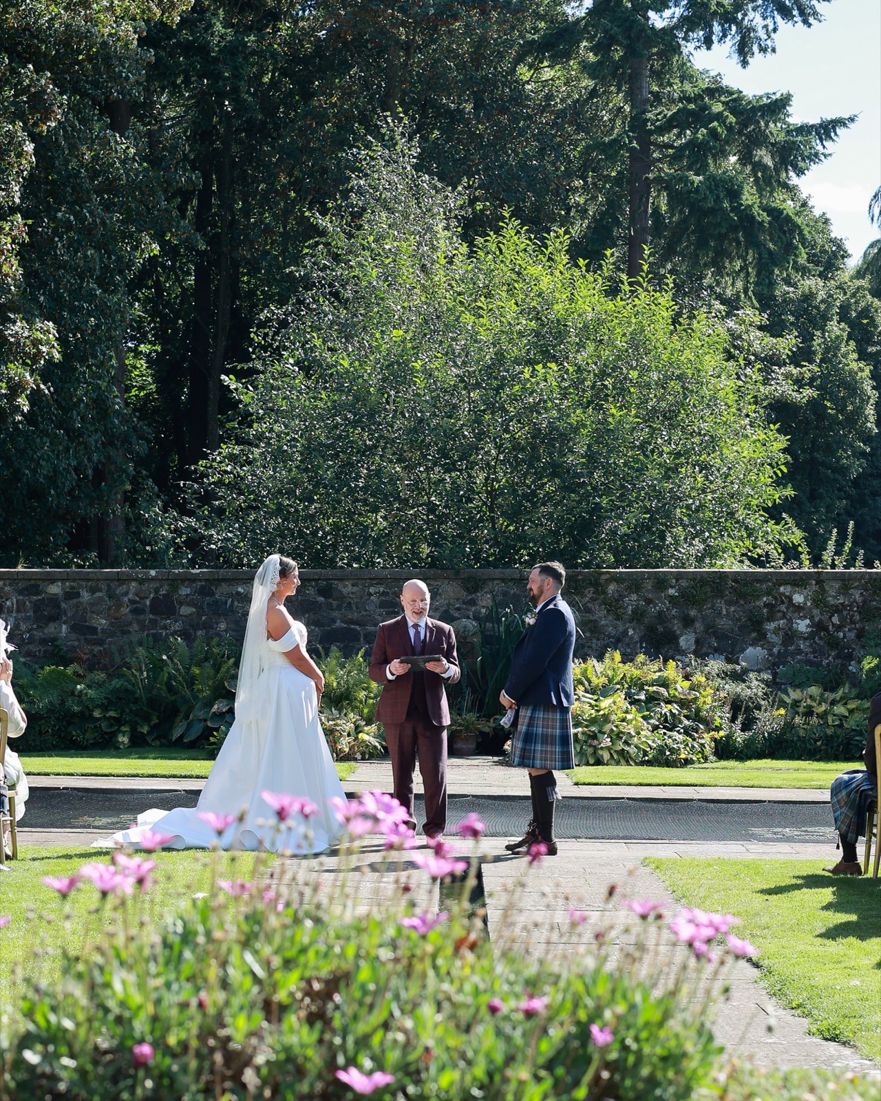 Wide shot of an outdoor wedding ceremony with seated guests and pink flowers in the foreground