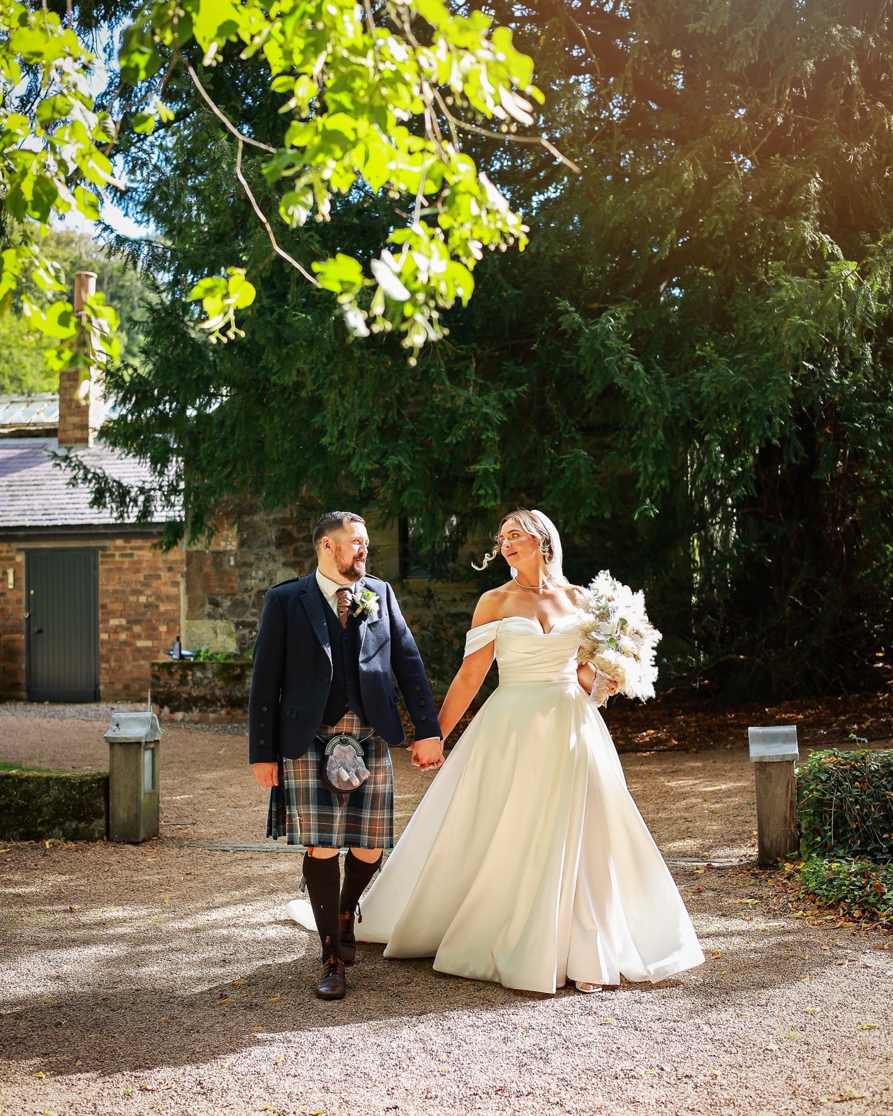 Bride and groom walking hand in hand along a gravel path, groom in a kilt