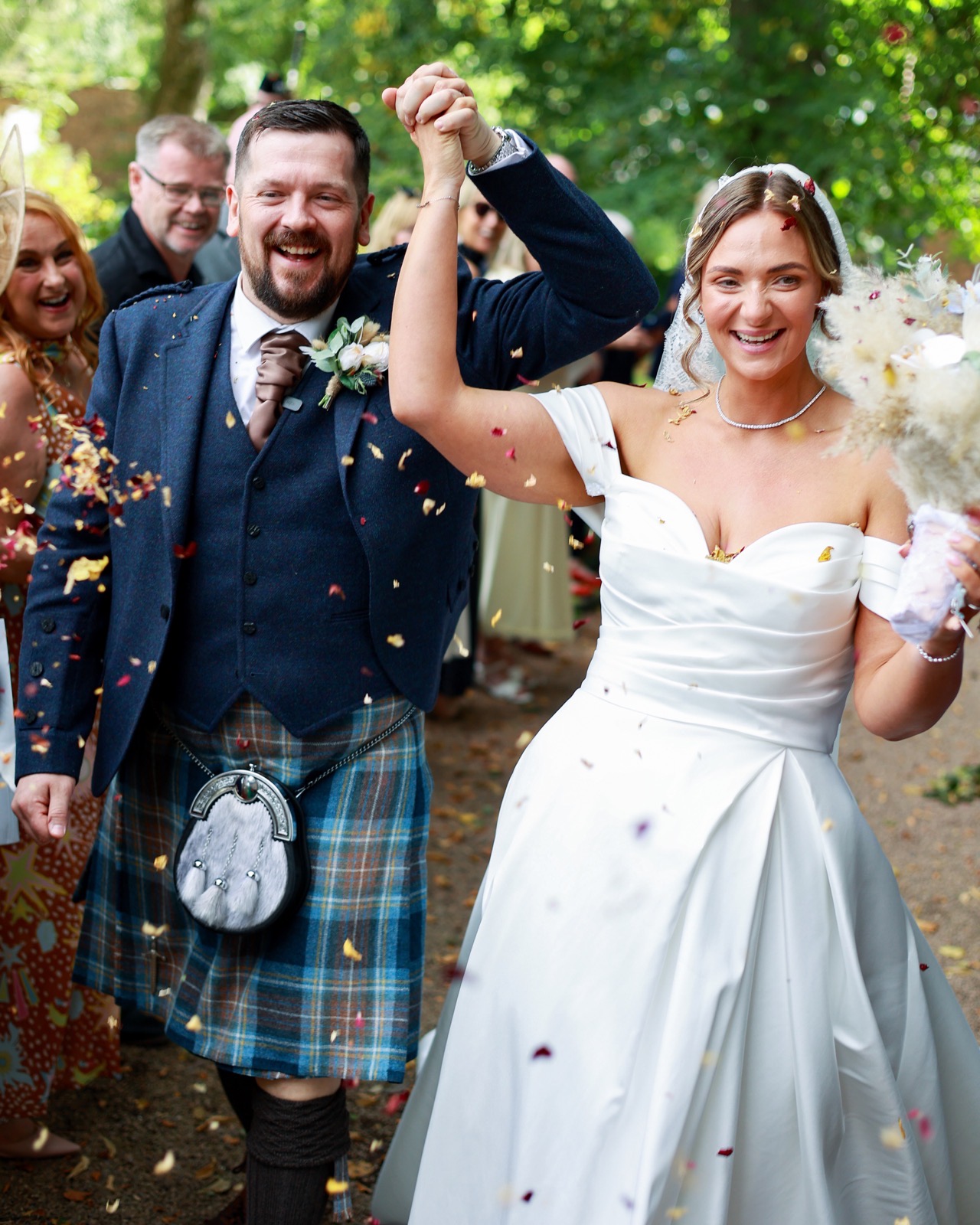 Bride and groom walking through a confetti shower with joined hands raised, guests cheering behind