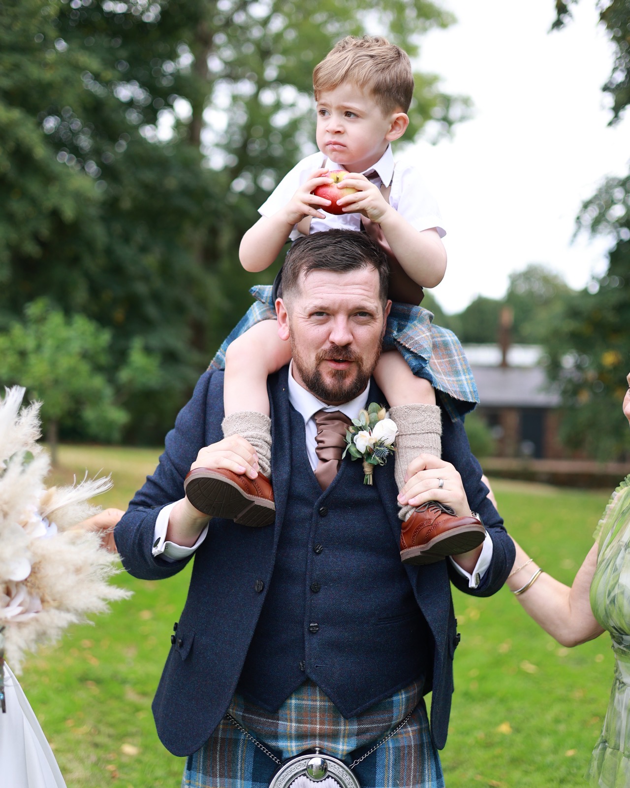 Groom carrying a young boy in a kilt on his shoulders while he eats an apple
