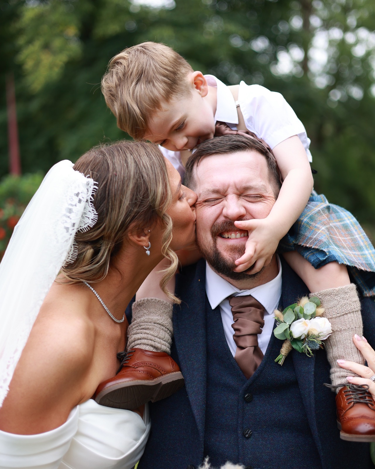 Young boy on the groom's shoulders leaning down to kiss his forehead, bride kissing the groom's cheek