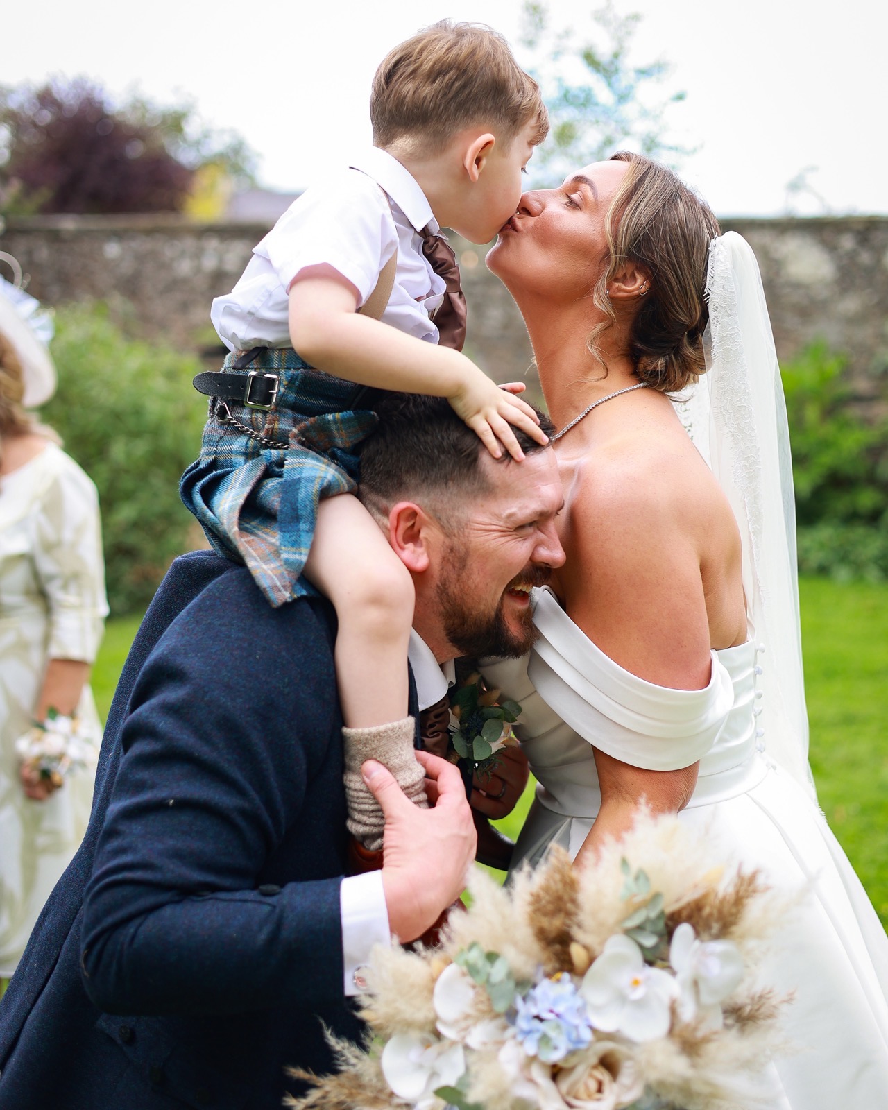 Young boy on the groom's shoulders leaning down to kiss the bride's cheek