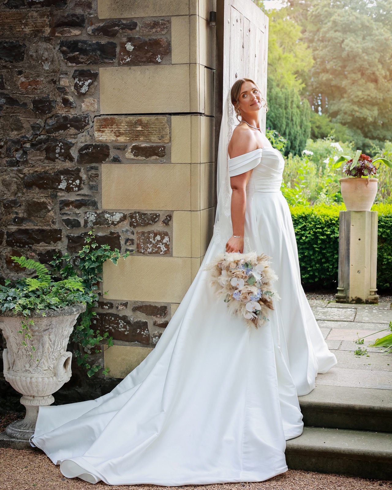 Bride standing alone by an old stone doorway, looking back over her shoulder with her bouquet