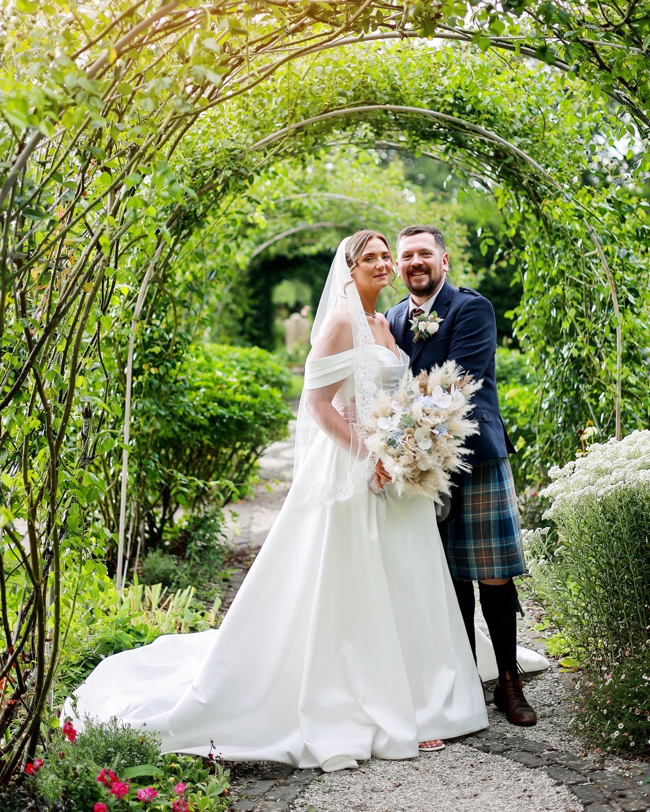 Bride and groom embracing under a green rose-covered archway tunnel