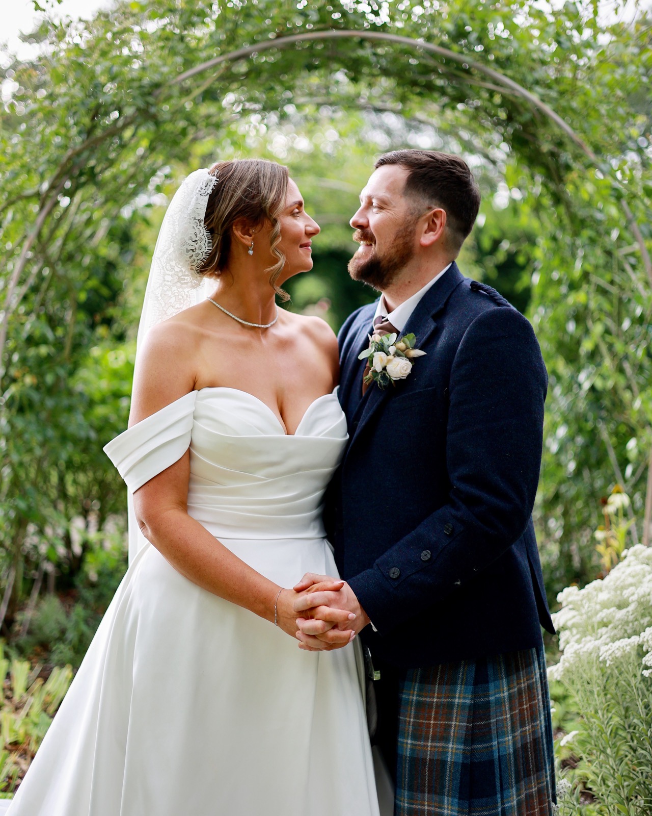 Close-up of the bride and groom holding hands and smiling, foreheads close, under a garden archway