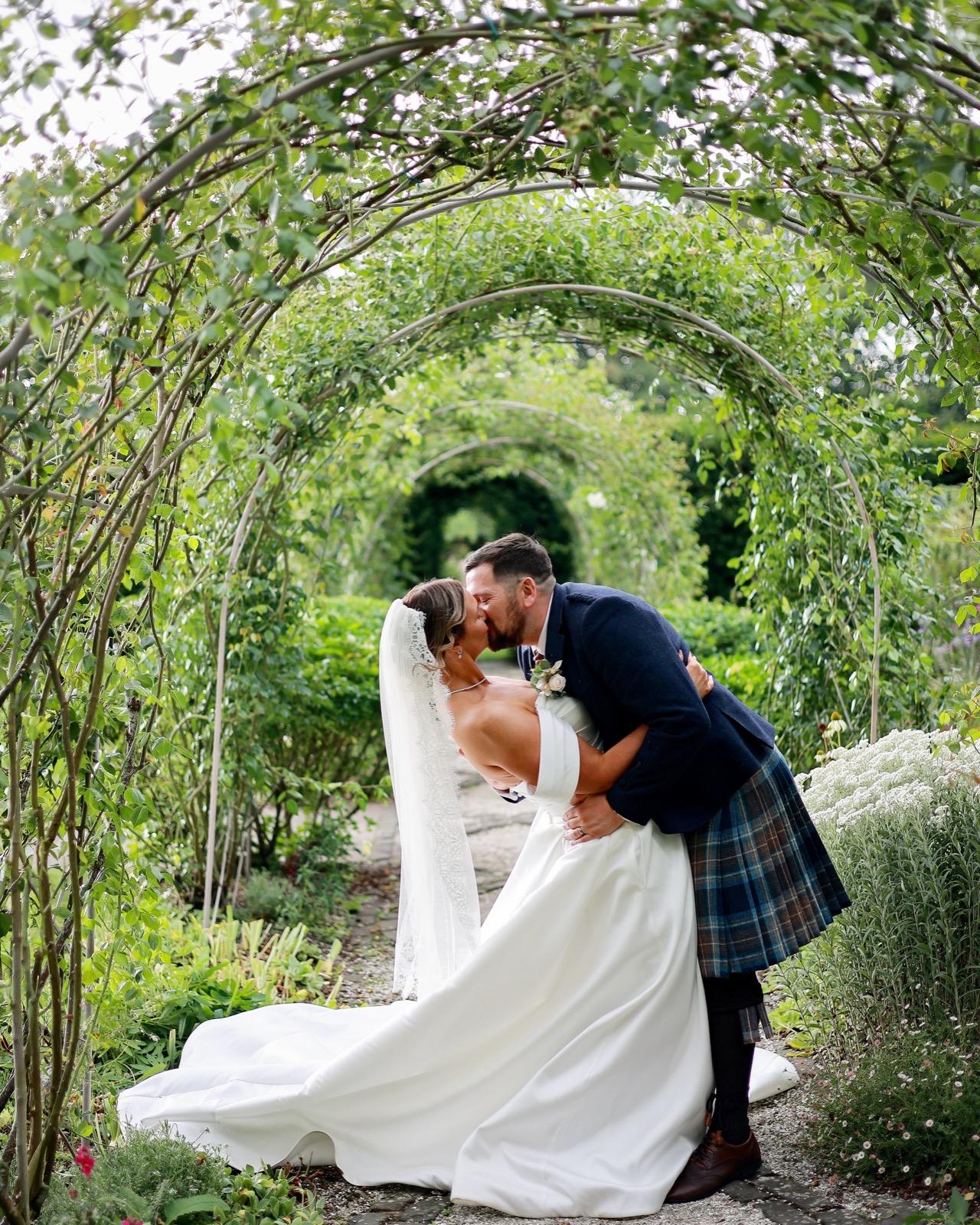 Groom dipping the bride slightly for a kiss under a rose-covered archway tunnel