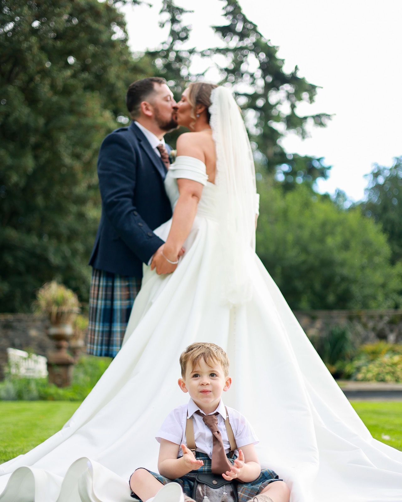 Bride and groom kissing with a young boy in a kilt sitting and clapping on the dress train in the foreground