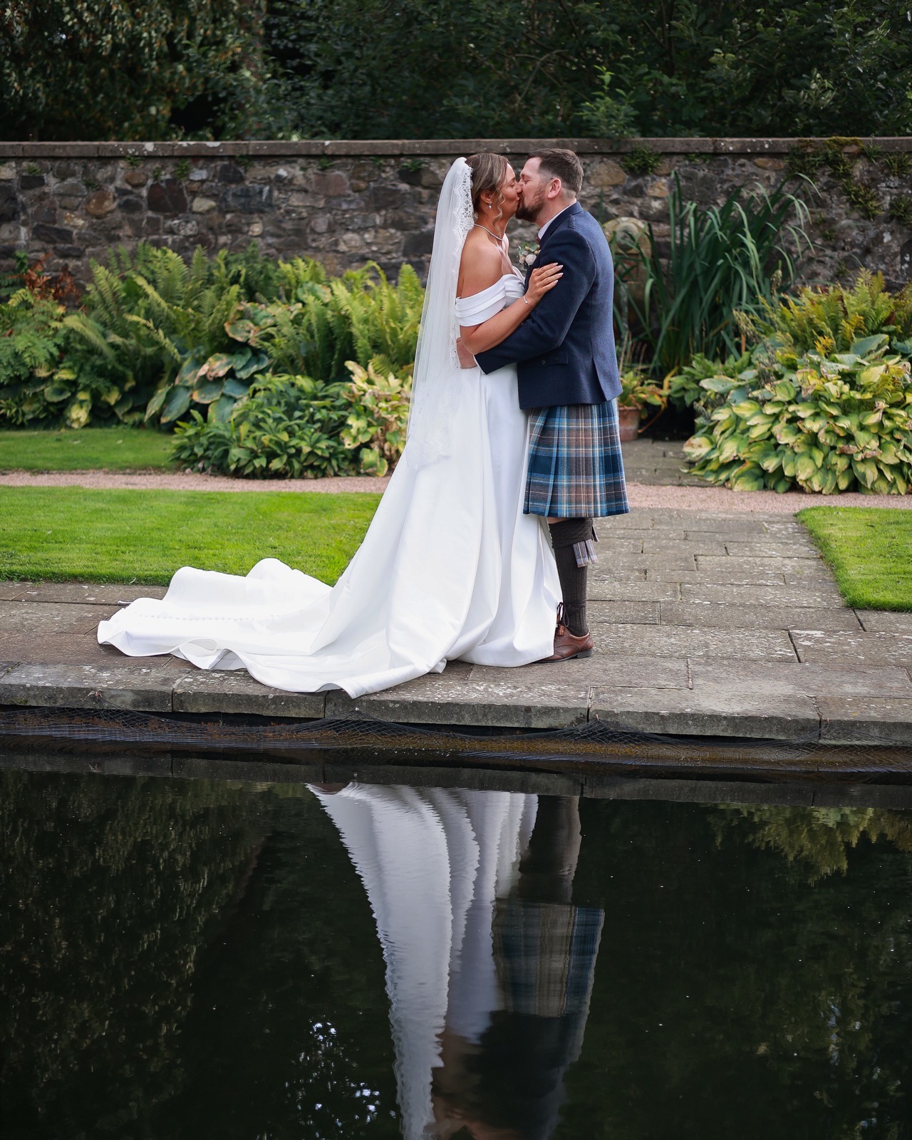 Bride and groom kissing beside a still reflecting pool, her dress train reflected in the water