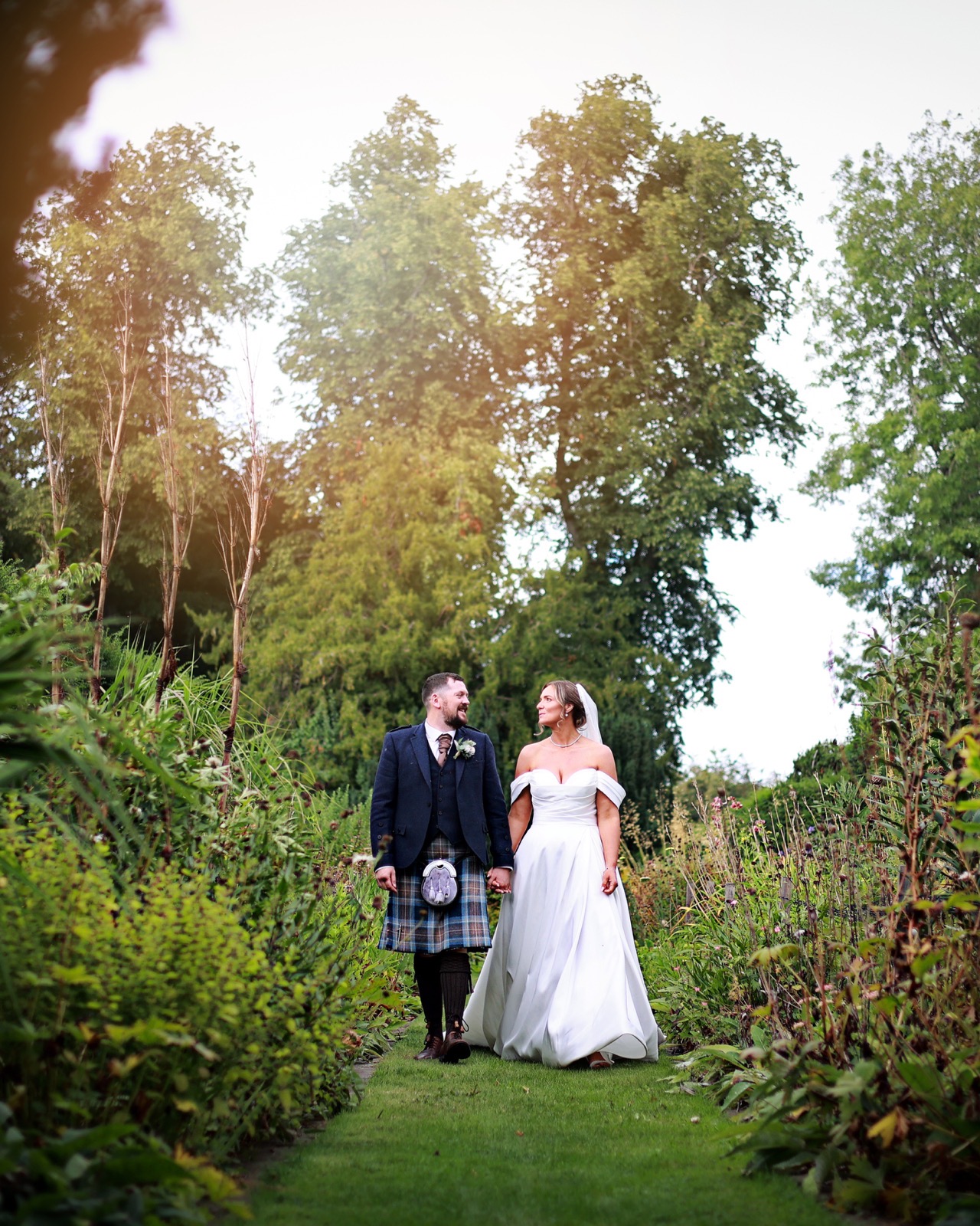 Bride and groom holding hands, walking together along a garden path lined with tall plants