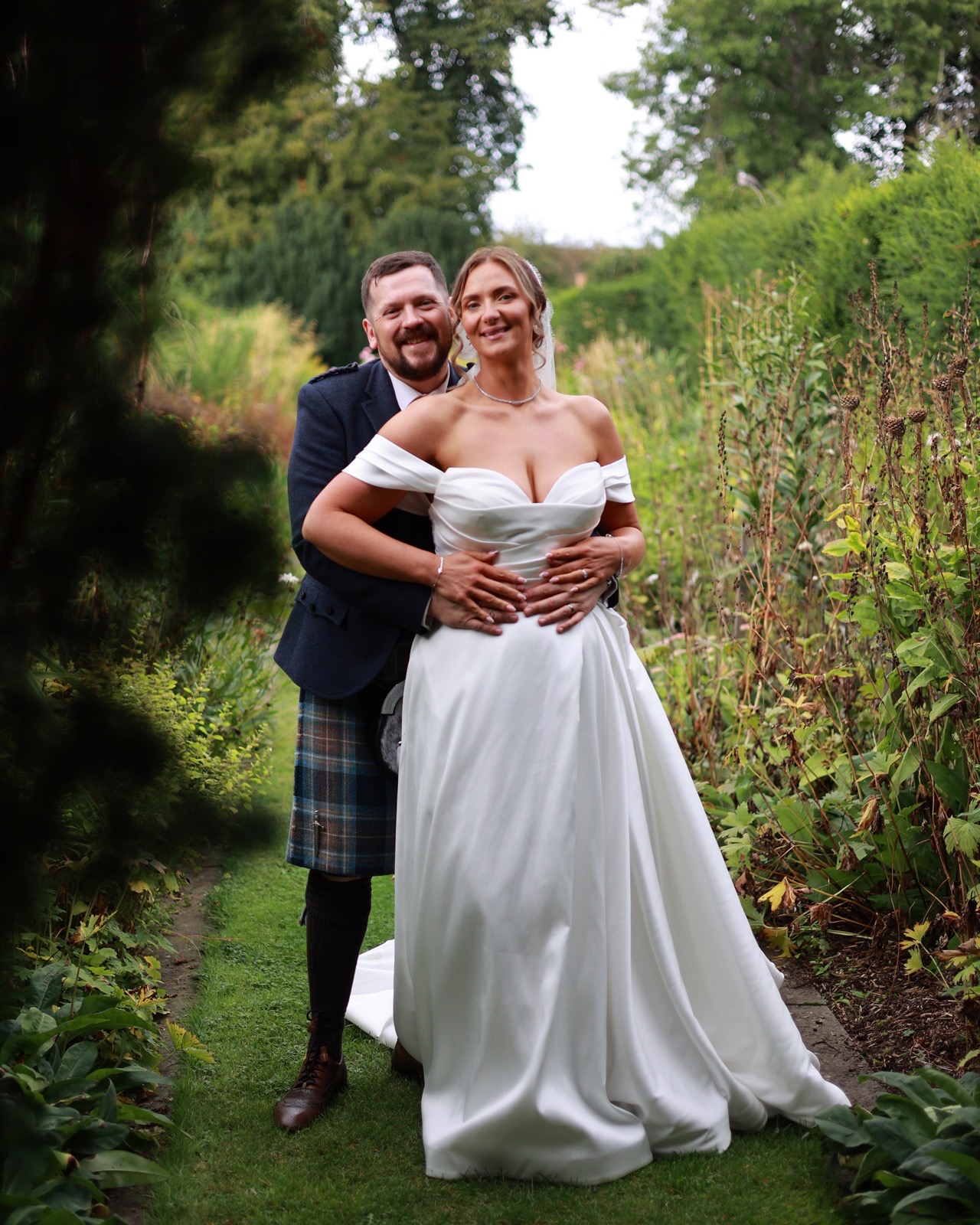 Groom hugging the bride from behind on a narrow garden path, both smiling