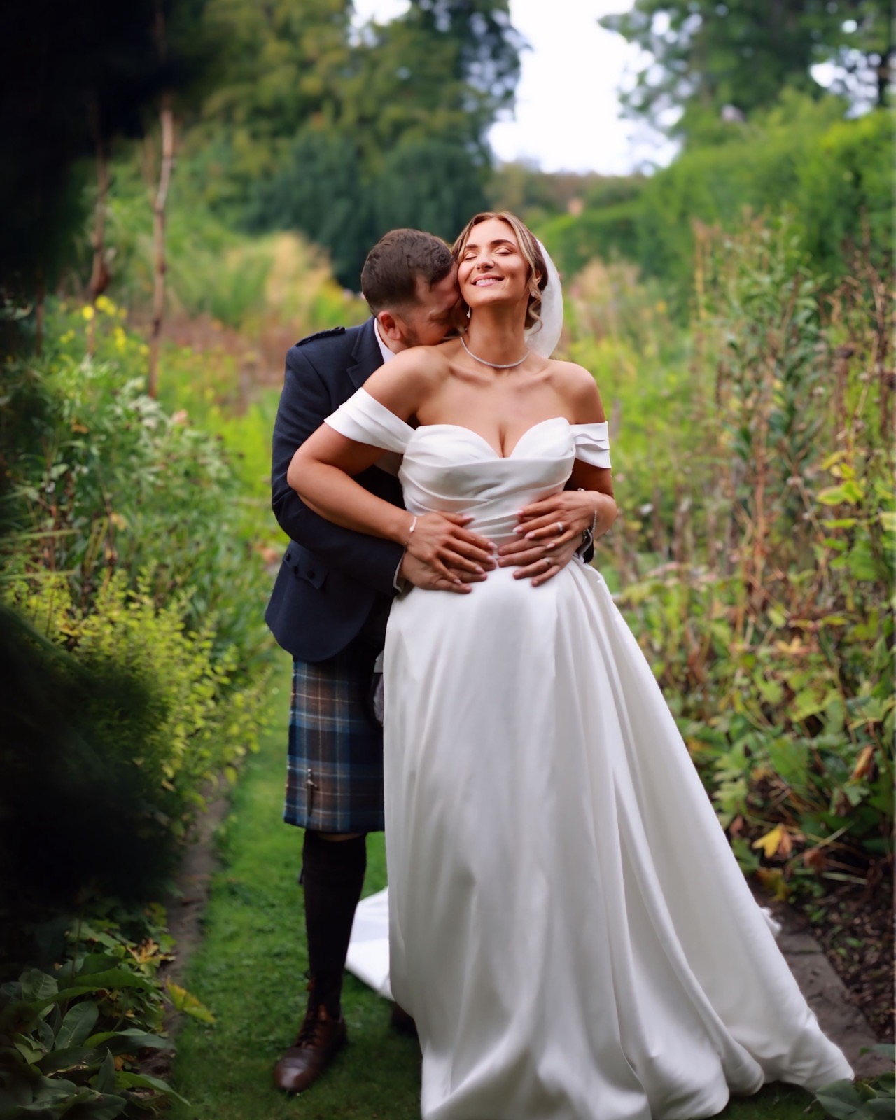 Groom kissing the bride's cheek from behind on a garden path, she smiles with her eyes closed