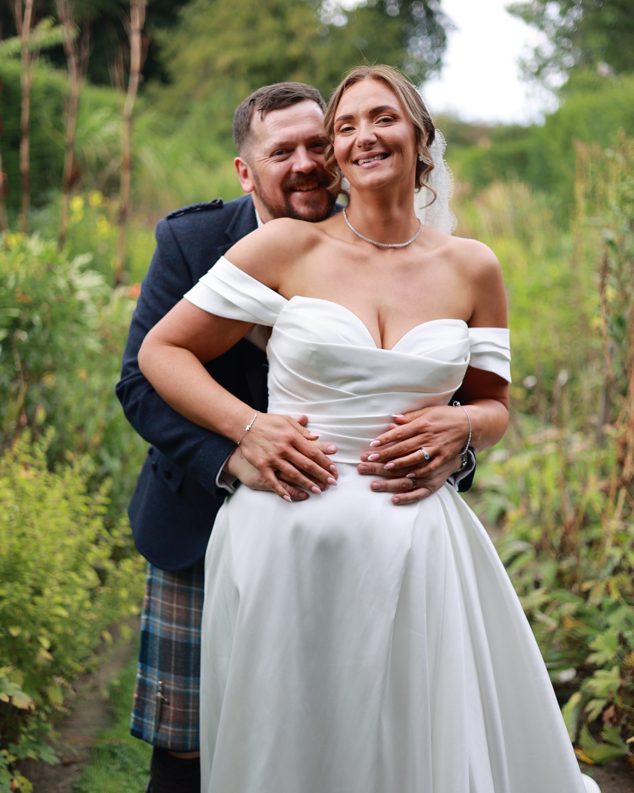 Groom hugging the bride from behind in a close garden portrait, both smiling