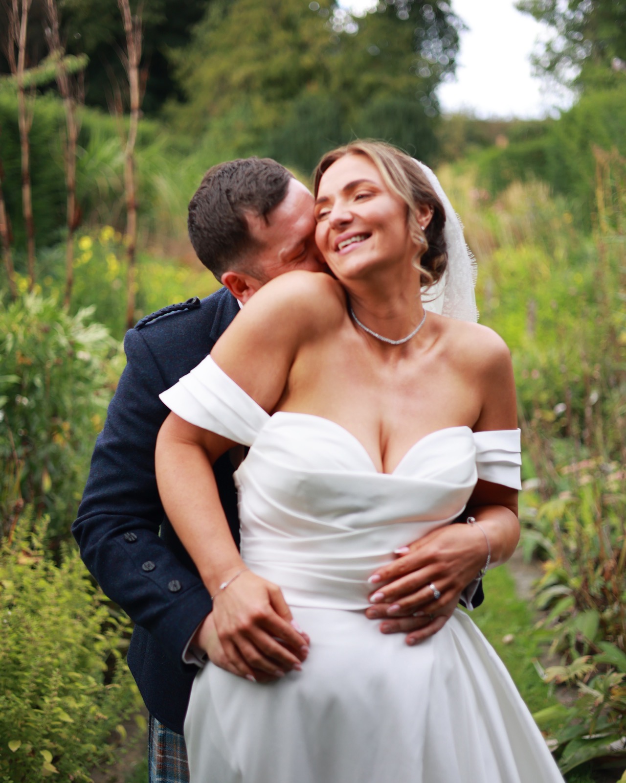 Groom kissing the bride's shoulder as she laughs, close garden portrait