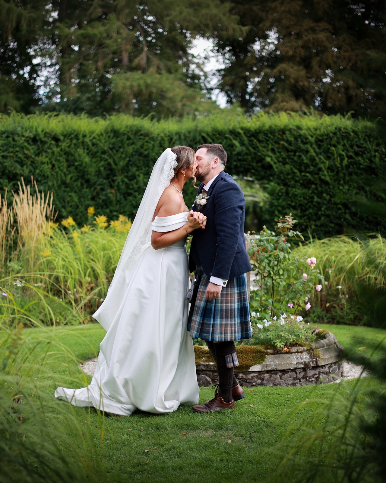 Bride and groom kissing beside a rose garden bed, holding hands