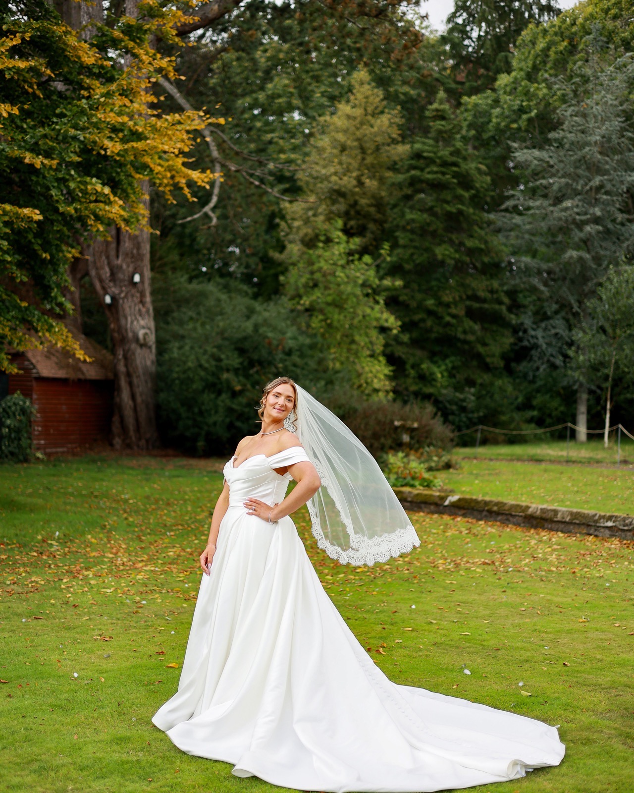 Bride standing with a hand on her hip, veil blown to the side, dress trailing across the lawn