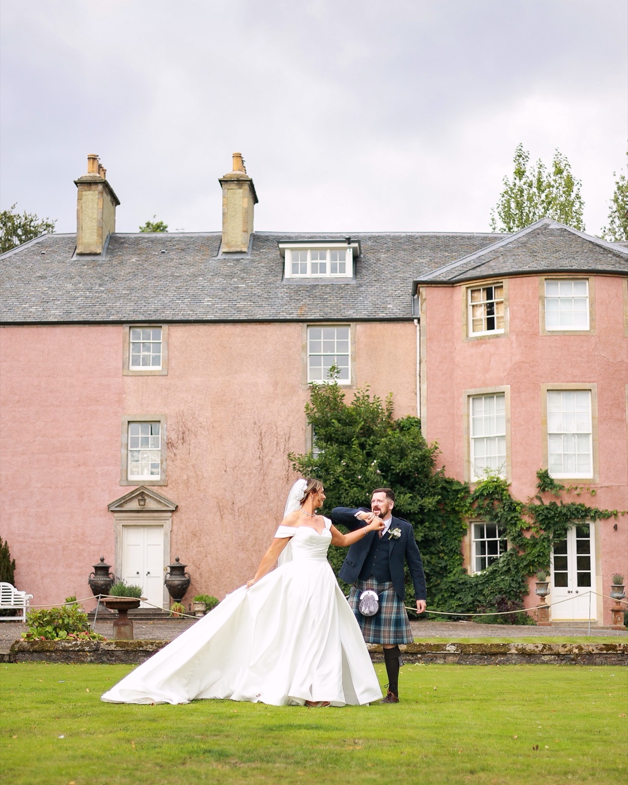 Bride and groom holding hands and walking across the lawn in front of a historic pink Scottish mansion house